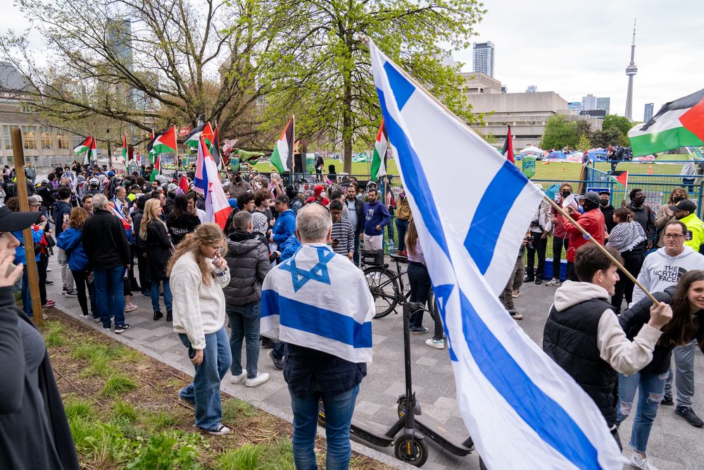 Pro-Israel supporters holding Israeli flags walks through crowd as pro-Palestinian and pro-Israeli protesters passionately express their views, with intense exchanges outside the student encampment occupying King's College Circle at the University of Toronto.
