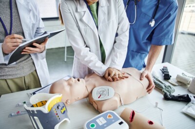 The image shows three individuals, likely medical professionals, practicing CPR on a medical training mannequin