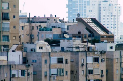 A dense cluster of older residential apartment buildings in Israel with weathered exteriors, rooftop satellite dishes, water tanks, and solar panels. The buildings have various air conditioning units and enclosed balconies. In the background, modern high-rise buildings are visible through a light haze, creating a contrast between old and new architecture.