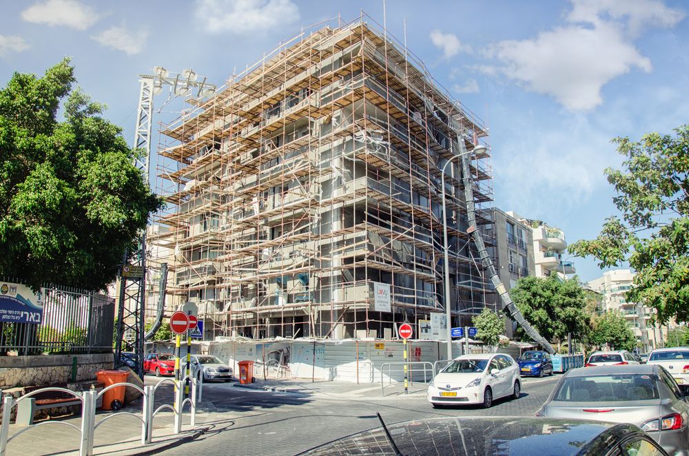 The image shows a residential (or possibly office) building undergoing renovation, renewal, or reconstruction, completely surrounded by scaffolding. There are street signs, parked cars, and regular urban traffic. The sky is clear, and there is plenty of greenery around. It appears to be a neighborhood in the midst of urban renewal.