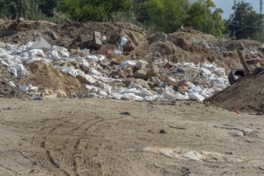 A pile of construction and demolition waste, including broken concrete, rocks, soil, white plastic bags, and discarded materials, dumped in an open area with tire tracks visible on the sandy ground.