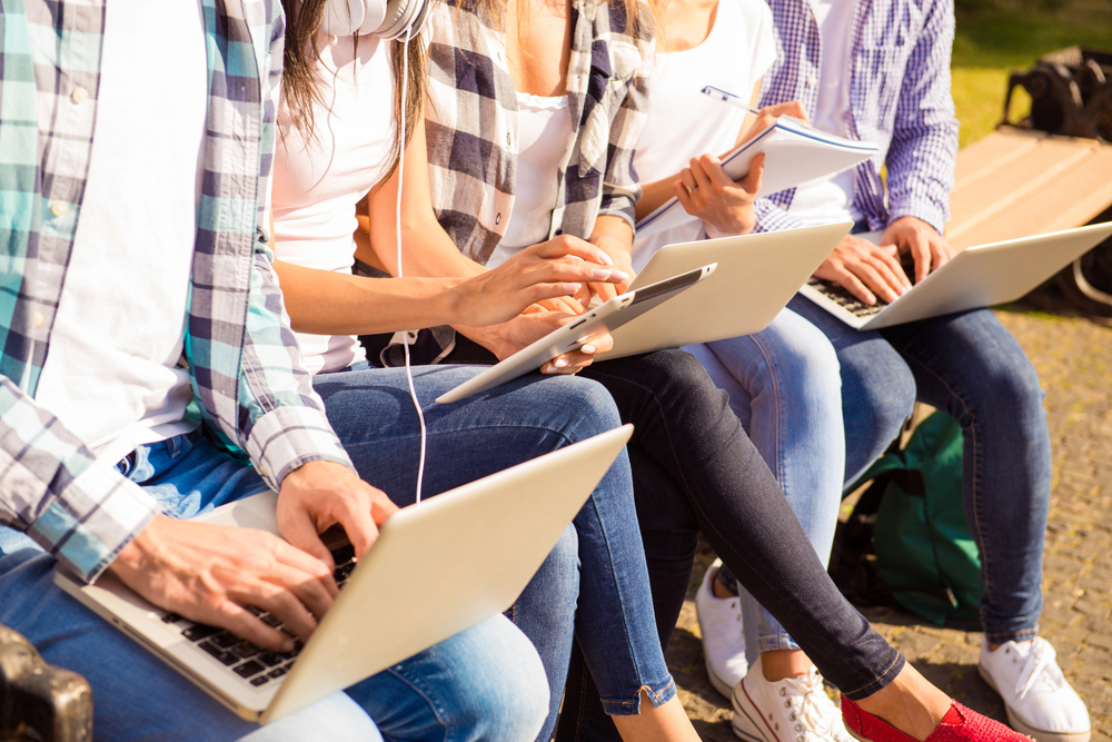 close-up photo of happy diverse classmates sitting on bench and study up with device